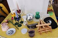 Multiple household items displayed on yellow tablecloth including vases, candle holders, disco ball, magnetic chess games, book stands, and travel bag.