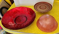Photo showing three glassware pieces - large red shallow bowl with floral swirl patterns, pink cake tray with cover, and red and gold ornate cake stand plate.