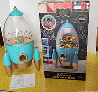 Rocket-shaped candy dispenser in blue and gold with clear top, original box and instruction manual on table.