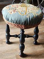 Front-side view of the Victorian round needlepoint footstool showing the floral basket embroidery on blue fabric and turned wooden black stained legs on a wooden floor.