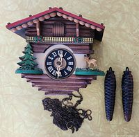 Front view of the vintage wooden cuckoo clock with a green pine tree and deer figurine beside the black clock face with Roman numerals, along with two pine cone-shaped weights and chains below.