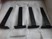 Four black metal L-shaped shelf brackets arranged side-by-side on white foam padding, showing vertical sides with mounting holes.