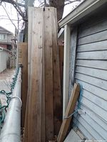 Photo showing a stack of multiple cedar boards leaning outdoors against a building, weathered surfaces visible.