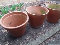 Three large clay planter pots arranged outdoors on soil with some leaves and garden plants nearby.