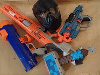 Four Nerf guns, a Minecraft toy hammer, and a black paintball mask arranged on a floor surface.