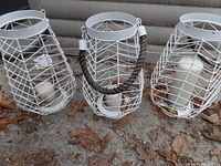 Three white metal wire outdoor candle holders with white pillar candles inside, arranged on a concrete surface with autumn leaves around them.