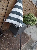 Photo of folded striped black and white patio umbrella leaning near a wooden fence and garden area.