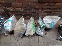 Photo showing five bags of pea gravel leaning against a brick wall on a concrete surface. Bags are mostly intact, with one or two slightly opened or dusty.
