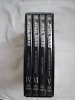 Star Wars Trilogy DVD collection showing four black DVD cases with white text: A New Hope (IV), Return of the Jedi (VI), Bonus Material, The Empire Strikes Back (V) held in a black slipcover box.