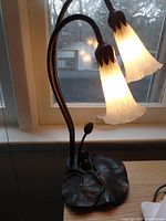 Full view of the lamp on a table near window, showing the bronze lily pad base and two flower shaped glass shades lit.