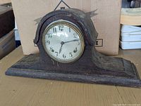 Antique solid wood mantle clock showing full side and front view on a wooden surface with cardboard box in background.