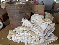 Photo showing the large antique Lard tin with faded advertising and rusty patina next to assorted vintage fabrics primarily lace and crochet in cream and off-white.