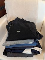 Stack of seven men's long sleeve casual shirts including a prominent black Lacoste shirt with green logo, folded and stacked on a chair.