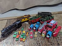 Wide shot showing group of metal trains and toy cars on carpet including Union Pacific tender and freight cars, black steam locomotive, caboose, blue Thomas trains, and various small cars and construction vehicles.