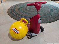 Red rider scooter toy and yellow bouncing ball shown together on floor with circular rug
