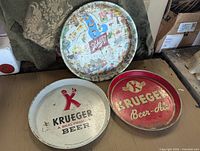 Three vintage round metal beer advertising trays with Krueger and Schlitz branding, laid on a cardboard surface including a light background with some surrounding items.