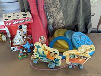 Overview of all toys including 101 Dalmatians figurine in box, Mickey Mouse bat, wooden pull toy train, Donald Duck figurine, plastic play discs in yellow and blue.