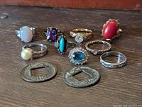 Photo showing full group of 8 vintage rings in various stone colors and 2 foreign coins with square holes on wooden surface.