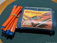 Photo of orange and blue plastic race track pieces bundled together next to blue Matchbox carry case with racing car graphics on top.