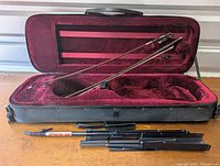 Open black violin case showing red velvet interior and two vintage violin bows inside.