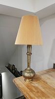 Full view of lamp showing brass base with intricate detailing and beige pleated fabric shade.