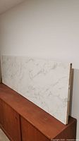 Photo of the faux marble patterned countertop panel resting on a wooden surface, showing the front face with white background and light gray marble veining.