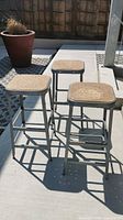 Three metal bar stools outdoors, showing full stools with shadow detail and a potted plant in the background.