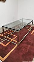 Full side view of the coffee table showing the thick glass top and stainless steel frame on a rug with geometric patterns.