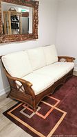 Front view of vintage rattan sofa with three white boucle cushions on geometric patterned rug in neutral indoor setting with decorative mirror on wall.