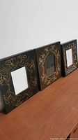 Three hand painted black wooden picture frames with gold decorative floral and scroll designs, placed side by side on wooden surface.