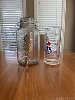 Photo showing two clear glass beverage containers against window blinds: a large pineapple drink dispenser with spigot and a glass pitcher with sports-related graphics.