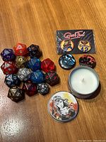 Photo showing the 14 D20 dice arranged on wood surface alongside four enamel pins and a Superwoman candle tin with the lid beside the candle.