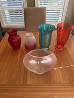 Five decorative glass vases displayed on wood table with window blinds in background, colors visible: red, pink ombré, blue, red/orange, and clear wide oval-shaped vase.