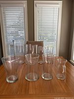 Front and back row clear glass vases arranged on wooden table near windows with blinds. The lot contains eight vases of varying heights and diameters, all transparent glass with smooth surfaces and no visible decoration.