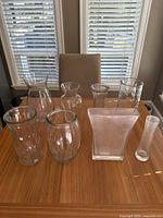 Photo showing all eight clear glass vases arranged on a wooden table, two rows, with various heights and shapes.