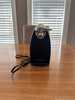 Front view of the black Hamilton Beach electric can opener showing its silver cutting mechanism and power cord looped beside it, placed on a wooden table.