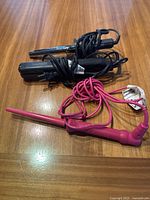 Three hair styling tools on wooden table including two curling irons with clamps and one pink curling wand, all cords wrapped.