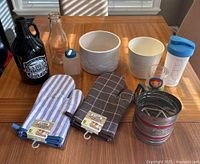 Full selection of kitchen items on a wooden table, including growler, milk bottle, pots, oven mitts, nesting cups, and flour sifter.