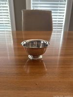 Front and side view of the small silver plated bowl on a wooden table with a chair and window blinds in the background.