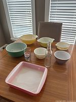 Seven Pyrex mixing bowls in different colors and sizes alongside a vintage Crush soda bottle and Molson Canadian glass on a wood table with bright daylight from windows.