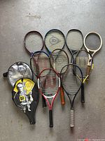 Full lot of nine tennis rackets arranged in two rows alongside two covers on a concrete floor.