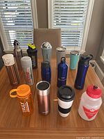 Front view of 11 assorted water bottles and travel mugs on a wooden table, showing brands and condition