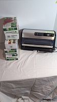 Photo of the vacuum sealer placed on a table alongside four boxes of FoodSaver vacuum sealer bags stacked upright.