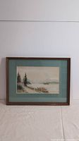 Front view of framed watercolor landscape painting featuring trees, shoreline, and mountains, with green mat and brown wooden frame.