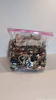 Clear resealable plastic bag filled with mixed jewelry components shown from front. Contains various beads, chains, and charms of different colors and materials.