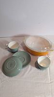 Photo shows a Wedgwood green glazed ceramic bowl upside down, two ceramic tea cups with saucers, and a yellow Pyrex ceramic casserole with glass lid.