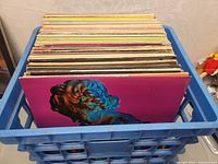 Blue crate filled with stacked vinyl record albums showing different colors and thicknesses.