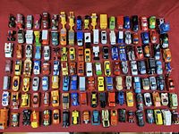 Top-down view of a large assortment of small die-cast toy cars arranged in rows on a red mat, showing various vehicle types including cars, trucks, and motorcycles in multiple colors.