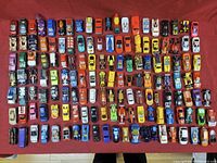 Top-down view of approximately 90 assorted die-cast toy cars arranged on a red cloth background. Variety of makes and styles visible.