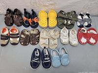 Photo showing 11 pairs of kids and infant shoes arranged neatly on floor including sandals, sneakers and slip-ons in multiple colors.